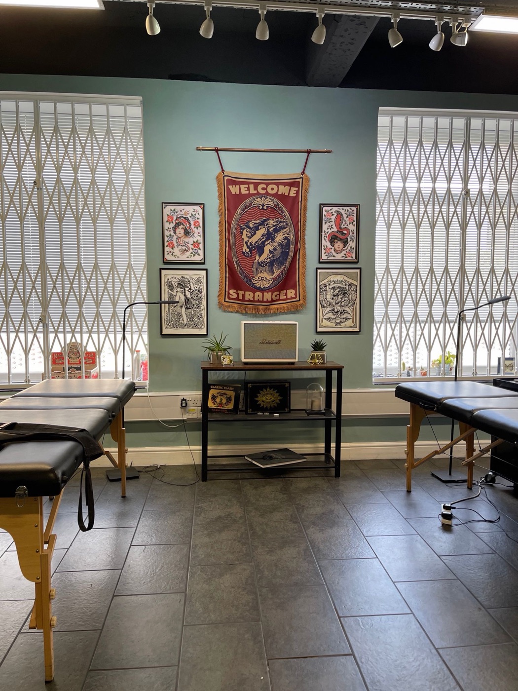 The Riverside Tattoo studio interior — sage-green walls with framed traditional tattoo flash, a Welcome Stranger banner and two clean tattoo benches by the windows